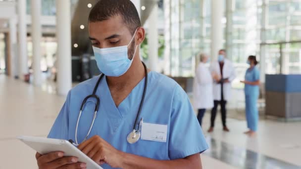 Portrait of young mixed race doctor using digital tablet while wearing safety surgical mask during Covid pandemic. Confident indian male nurse working on digital tablet at hospital wearing face mask against Covid-19 while looking at camera. - Imágenes, Vídeo