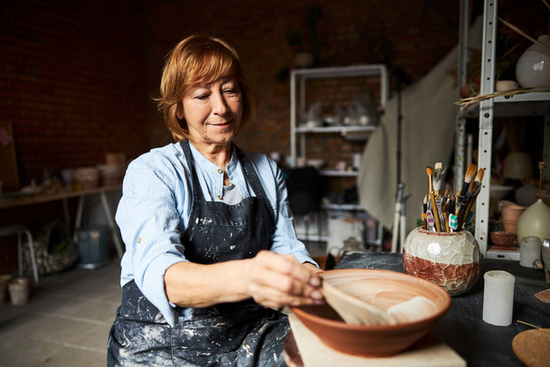 Nice old woman ceramist painting clay bowl in workshop - Photo, Image