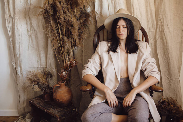 Stylish fashionable woman in beige suit and hat posing on wooden chair in bohemian style room with dried grass and flowers in vase. Young attractive female sitting in atmospheric boho interior - Foto, imagen