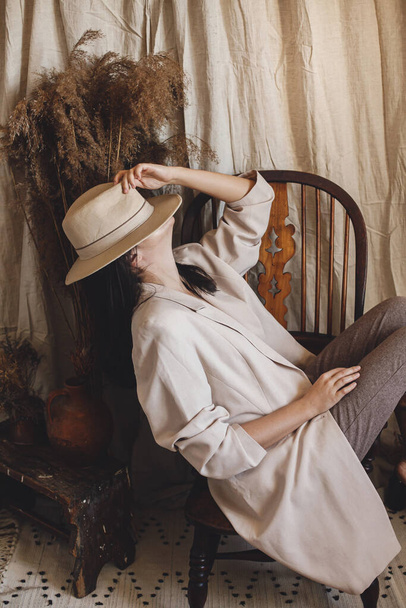 Mooie stijlvolle vrouw in pak zittend op retro houten stoel op de achtergrond Boheemse stijl kamer met gedroogd gras en bloemen in vaas. Jonge modieuze vrouw poseren in pastel boho interieur - Foto, afbeelding