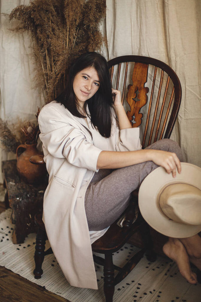 Beautiful stylish woman in suit sitting on retro wooden chair on background bohemian style room with dried grass and flowers in vase. Young fashionable female posing in pastel boho interior - Foto, Imagem