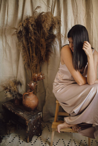 Elegante mujer de moda en vestido de seda posando sensualmente en habitación de estilo bohemio con hierba seca y flores sobre fondo de tela beige. Joven atractiva hembra sentada en el interior boho atmosférico - Foto, imagen