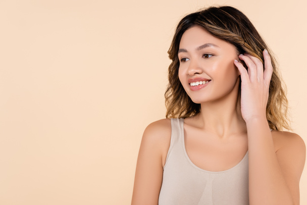 nuori aasialainen nainen vahvistamisesta hiukset ja hymyillen eristetty beige - Valokuva, kuva