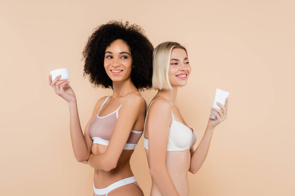 smiling interracial women holding cosmetic cream while standing back to back isolated on beige - Foto, afbeelding