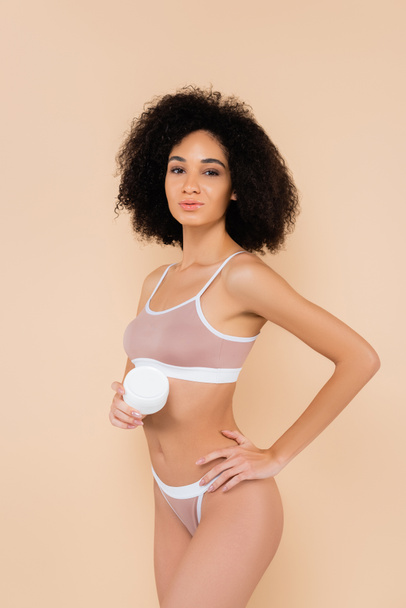 african american woman holding cosmetic cream while posing with hand on hip isolated on beige - Foto, immagini