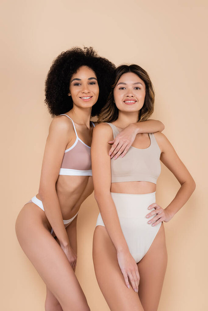 african american woman hugging asian friend while posing in underwear isolated on beige - Zdjęcie, obraz