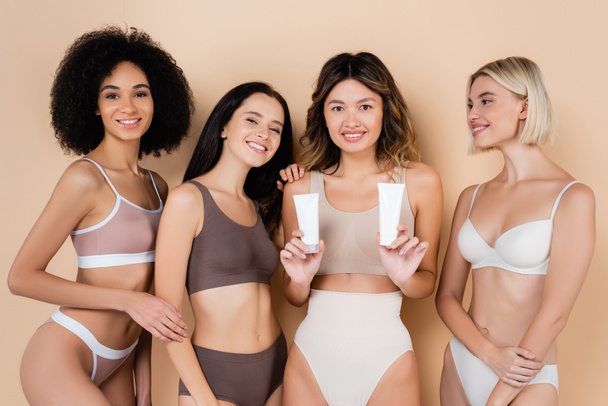 asian woman holding tubes of body cream near multicultural friends in underwear on beige - Photo, Image