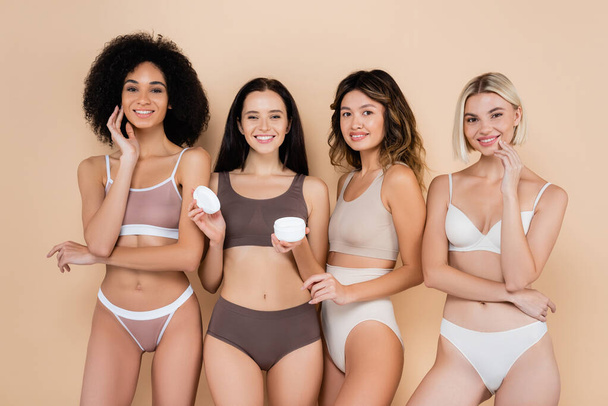 joven mujer sosteniendo crema cosmética cerca bastante interracial amigos en beige - Foto, Imagen