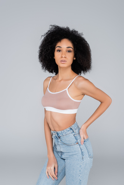 african american woman in jeans and bra posing with hand in back pocket isolated on grey - Фото, изображение