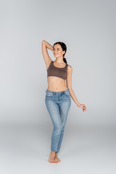 smiling woman in jeans and bra looking away while posing on grey - Valokuva, kuva
