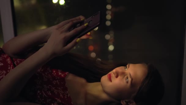 girl holds phone and looks at it at window against background of night city. - Footage, Video
