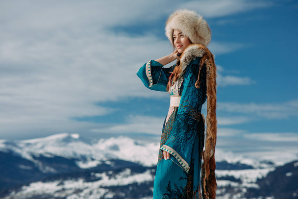 Mujer joven está vestida con sombrero de piel, vestido y cuello de zorro contra el hermoso paisaje de los Cárpatos nevados montañas y el cielo. - Foto, Imagen