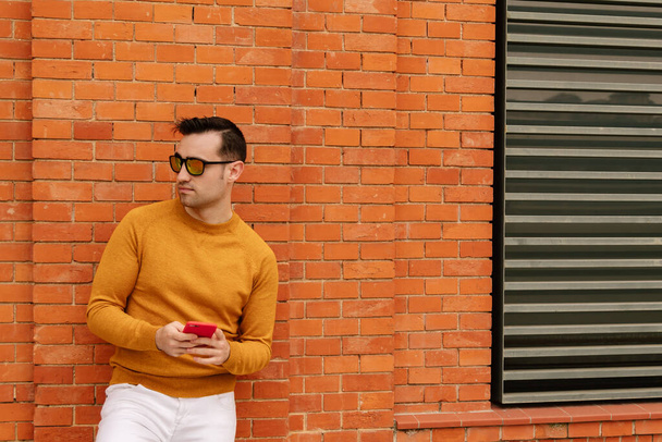 Attrayant et jeune homme aux lunettes de soleil orange utilisant son smartphone, vêtu de vêtements jaunes et de murs en briques. concept mode de vie. - Photo, image