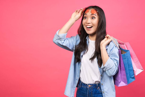 Photo of beautiful emotional surprised young cute asian woman posing isolated on pink wall background in denim clothes, holding shopping bags. Concept for discounts, sale, black friday. Copy space. - Φωτογραφία, εικόνα