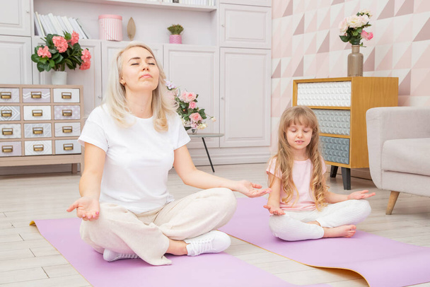 Full length front view smiling blond mother on yoga mat with cute playful little preschool daughter do various exercises. Happy mommy practicing yoga exercised with small child. - Фото, изображение