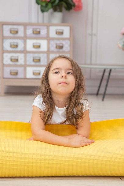 Full length front view smiling cute playful little preschool girl sitting on yoga mat looking at the camera. Happy child practicing yoga exercised at home. - Fotó, kép