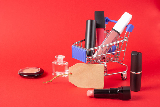A set of lipstick cosmetics and lip glosses, powder, eyeshadow and a shopping trolley on a bright red background. The concept of buying cosmetics, online shopping, holiday - Foto, immagini