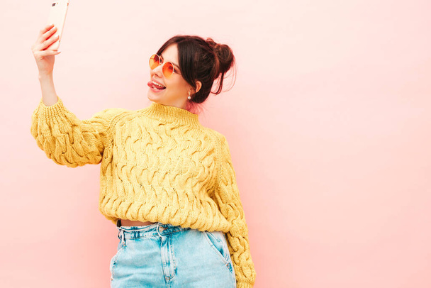 Joven hermosa hembra sonriente en moda verano suéter hipster amarillo y jeans. Sexy mujer despreocupada posando cerca de la pared rosa en el estudio. Modelo positivo que se divierte en gafas de sol. Tomar selfie - Foto, imagen