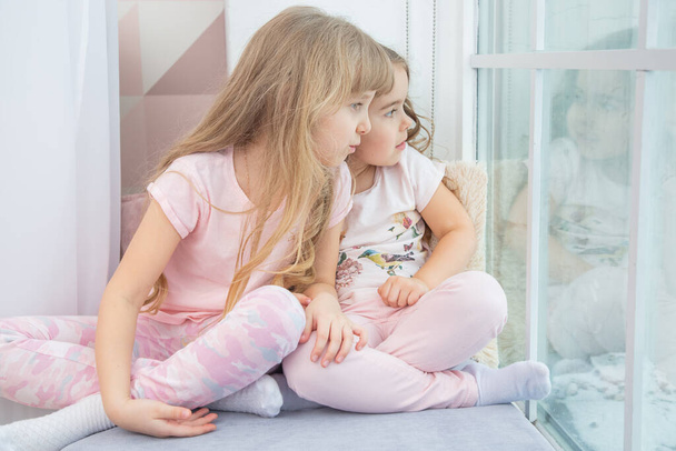 Cute little siblings sitting on windowsill at home looks out window to snow, portrait of pretty little girls on room sill in winter. Adorable pensive child in pink, cute toddler during contemplation. - Photo, image