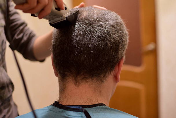 Ein erwachsener Mann wird mit einem Haarschneider beschnitten und sitzt mit dem Rücken zur Kamera. Vielleicht zu Hause im Sperrbezirk - Foto, Bild