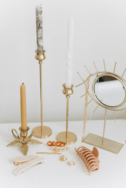 Boho accessories, interior details. Modern golden jewellery, hair clips on white table with vintage candles and boho mirror. Stylish gold ring, chain necklace, earrings, hairpins and barrettes. - Photo, Image