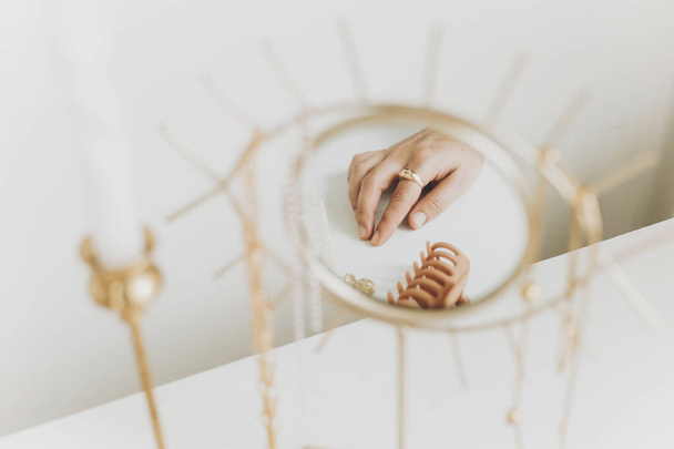 Main féminine avec bague dorée moderne et boucles d'oreilles réfléchies dans un miroir boho sur une table blanche avec des bougies vintage. Bague élégante en or, collier, épingles à cheveux. Bijoux Boho - Photo, image