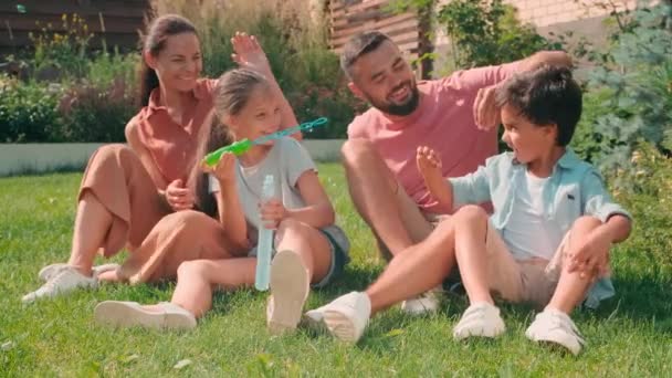 Полный снимок современной семьи с двумя детьми, проводящей летний день на заднем дворе, дети надувают мыльные пузыри и веселятся, пока над ними смеются родители - Кадры, видео