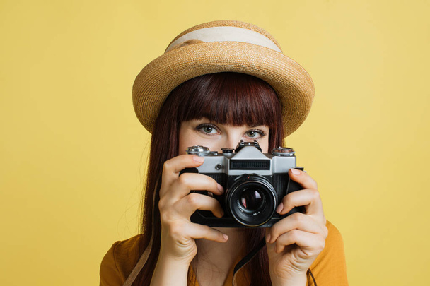 Hobby, taking photos, travel concept. Gorgeous young woman in hat, photographer amateur, smiling and taking picture on old vintage camera, on colorful yellow studio background - 写真・画像