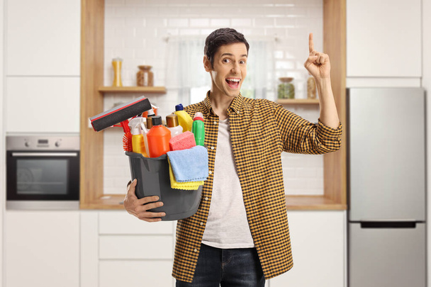 Young man with a bucket full of cleaning supplies in a kitchen pointing up and looking at camera - Foto, Imagem