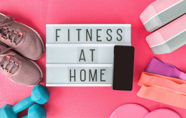 Fitness at home sign with mobile phone on pink yoga mat, running shoes, kettlebell weight and dumbells resistance bands and sliders for pilates online class. Exercise indoors for women. - Фото, зображення