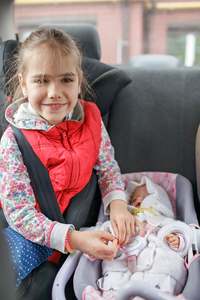 Dziewczyna siedzi w fotelikach samochodowych i bawi się lalką, która również w foteliku samochodowym, bezpieczeństwo transportu dzieci - Zdjęcie, obraz