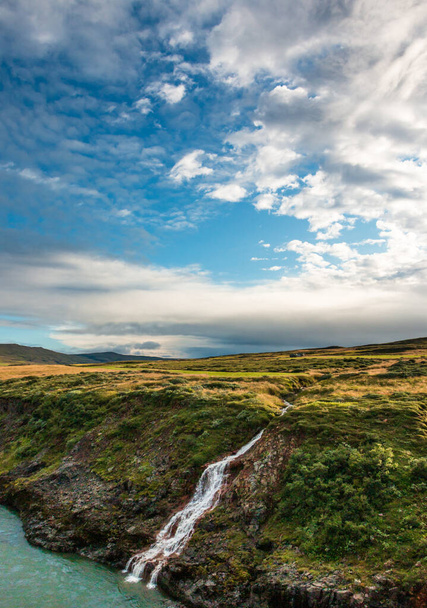 Cachoeira sobre a margem íngreme do rio que contribui para o rio turquesa colecionada sob o céu azul antes do pôr-do-sol, Islândia - Foto, Imagem