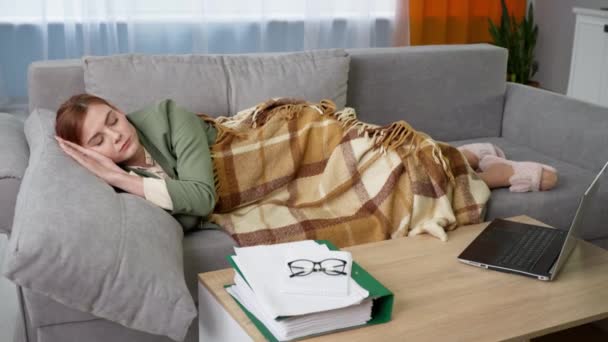 nainen takissa nukkuu peiton peitossa makaa sohvalla lähellä pöytää kannettavan tietokoneen ja asiakirjojen aikana etätyön karanteenissa - Materiaali, video