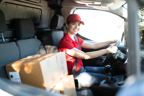 Mujer partera alegre conduciendo una furgoneta y haciendo contacto visual. mensajero femenino caucásico sentado detrás del volante y empezando a hacer entregas de paquetes - Foto, imagen