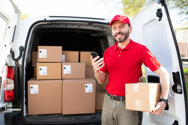 Portrait of a happy hispanic man smiling and making eye contact while holding a smartphone and a small package outside a delivery van - Photo, Image
