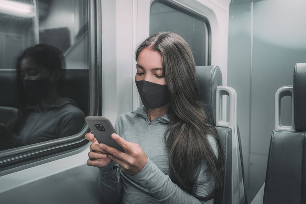 Public transport commute woman commuter at night wearing face mask riding subway using mobile phone. Asian passenger with face coronavirus covering on train commute ride. - Foto, afbeelding
