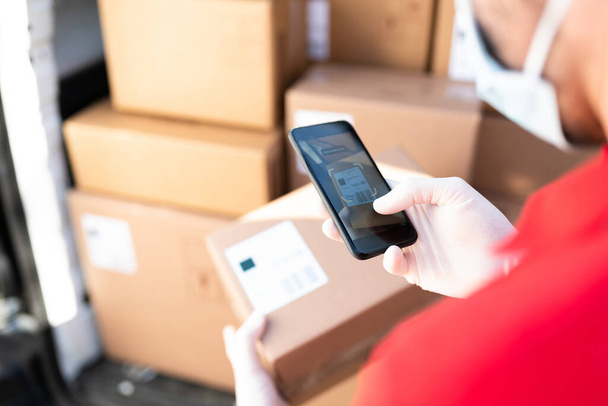 Close up of a male worker and delivery person with a face mask holding a smartphone and scanning the barcode of a shipping label to deliver a package - Fotoğraf, Görsel