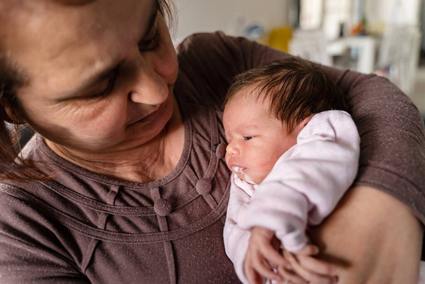 Ανώτερη καυκάσια γιαγιά κατέχουν νεογέννητο βρέφος κορίτσι εμετό γάλα - μωρό υπερσίτιση εμετό γάλα στα χέρια της γιαγιάς της στο σπίτι - νέα ζωή νοσηλευτική και μεγαλώνοντας έννοια - Φωτογραφία, εικόνα