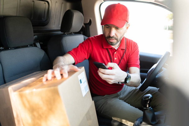Correo masculino hispano escaneando el código de barras de una etiqueta de envío de un paquete mientras está sentado al volante de una furgoneta de entrega antes de entregar el paquete - Foto, imagen