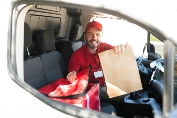 Retrato de um homem sorrindo em seus 30 anos entregando um saco marrom com almoço ou jantar. Entrega homem dirigindo uma van branca e fazendo um serviço de entrega em domicílio de comida de takeout - Foto, Imagem