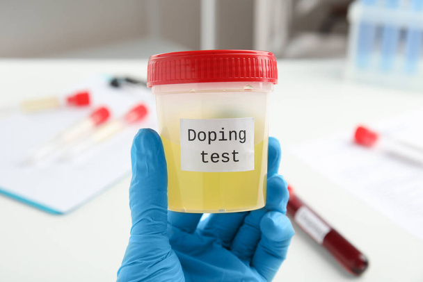 Doctor holding jar with urine sample indoors, closeup. Doping control - 写真・画像