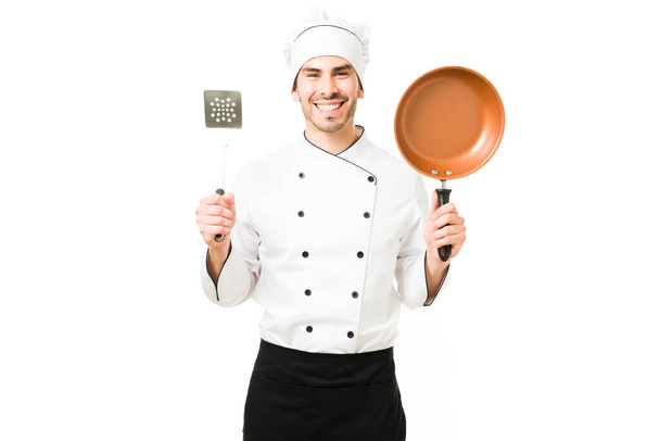 帽子をかぶった白い制服を着た幸せな若い男が、高級レストランでシェフとして働いています。白い背景の前に鍋とへらを持ったおいしそうな料理 - 写真・画像