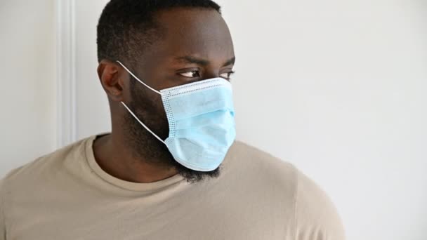 Afro-Américain en vêtements décontractés se tient à l'intérieur avec un masque médical de protection sur le visage et regarde dans la caméra tout en restant isolé à la maison. Précautions de quarantaine, concept de santé - Séquence, vidéo