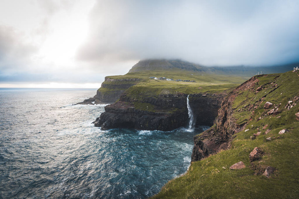 Aerial drone view of Gasadalur village and Mulafossur its iconic waterfall, Vagar, Faroe Islands, Denmark. Rough see in the north atlantic ocean. Lush greens during summer. - Фото, изображение