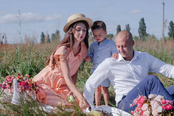 glückliche Familie bei einem Picknick auf dem Feld. Mann, Frau und kleiner Junge sitzen auf einer Decke mit Blumen, Wein, Baguette und Früchten in der Nähe. romantisches Date. Kindererziehung. Mutter, Vater und Sohn zusammen - Foto, Bild