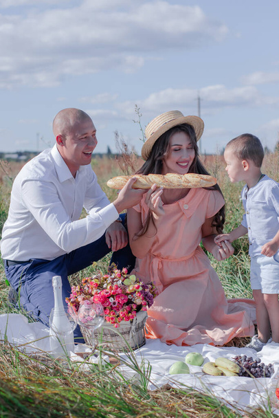 счастливая семья на пикнике в поле. муж, жена и маленький мальчик сидят на одеяле и едят багет. цветы, вино и фрукты поблизости. воспитание. мать, отец и сын вместе - Фото, изображение