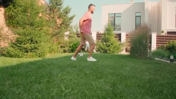 slow-motion low-angle shot van moderne jonge volwassen man voetballen met schattige 8-jarige zoon op zonnige zomerdag in de achtertuin - Video