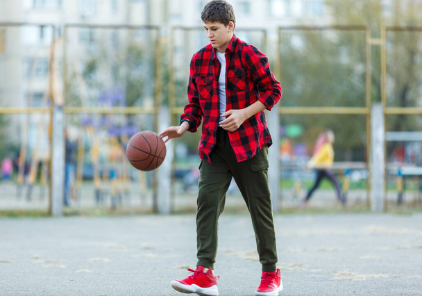 Ένα χαριτωμένο αγοράκι παίζει μπάσκετ σε μια παιδική χαρά. Teenager σε κόκκινο φανελένιο πουκάμισο με πορτοκαλί μπάλα μπάσκετ έξω. Χόμπι, ενεργός τρόπος ζωής, αθλητική δραστηριότητα για παιδιά. - Φωτογραφία, εικόνα