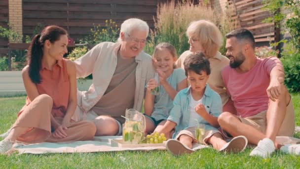 Slow-motion full family portrét nadšené elementární sourozenci tráví letní den se svými rodiči a prarodiči chlazení na trávníku v zahradě chatování a pití limonády - Záběry, video