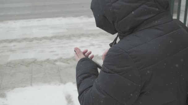 Esik a hó, egy nő meleg kabátban az út szélén taxit hív a telefonján keresztül. - Felvétel, videó